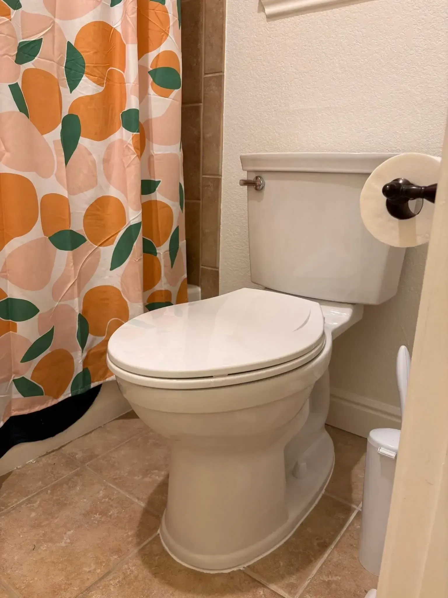 A white toilet in a bathroom with a peach-patterned shower curtain and a roll of toilet paper on a wall mount.