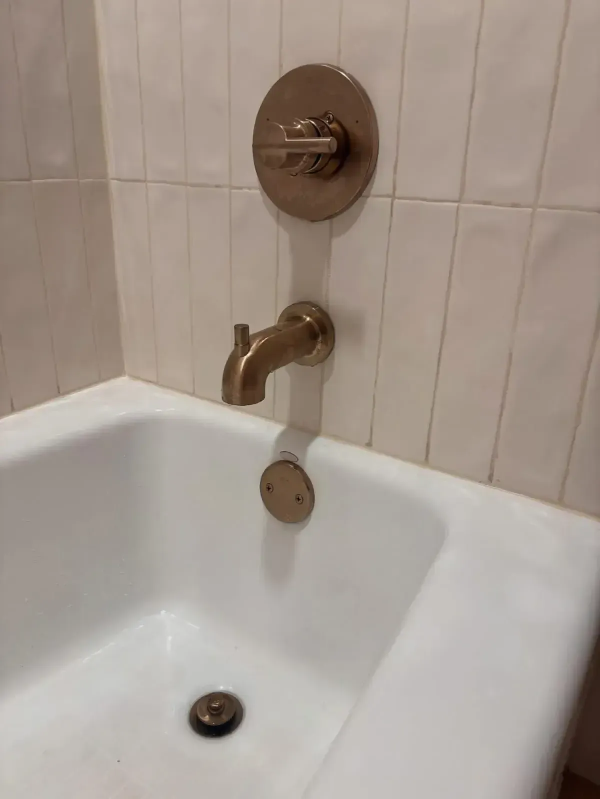 A brass-toned bathtub faucet and control handle on a wall with white vertical tiles above a white porcelain tub.
