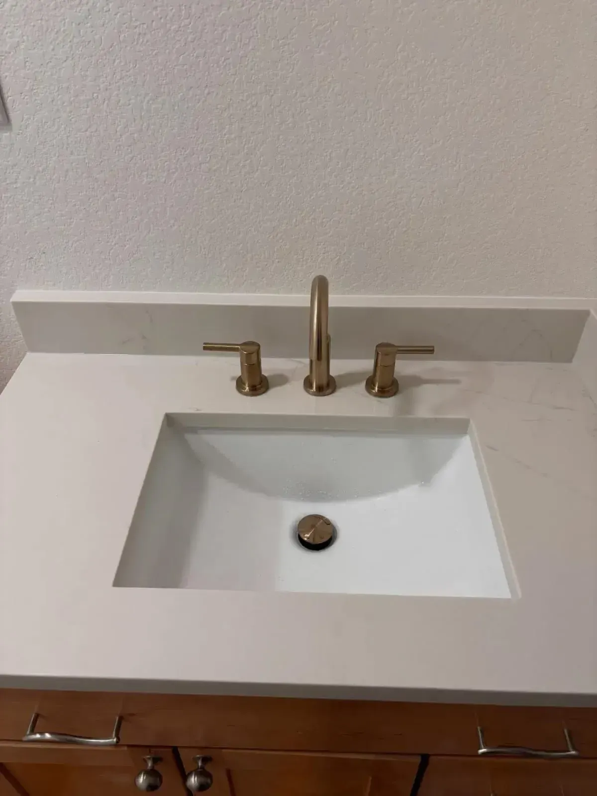 A bathroom vanity with a white rectangular sink, a gold-toned faucet, and a light-colored countertop.