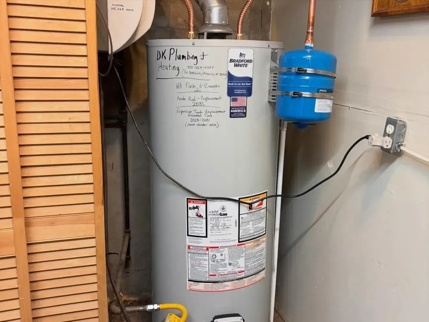 A grey electric water heater with a blue expansion tank mounted on a wall next to it.