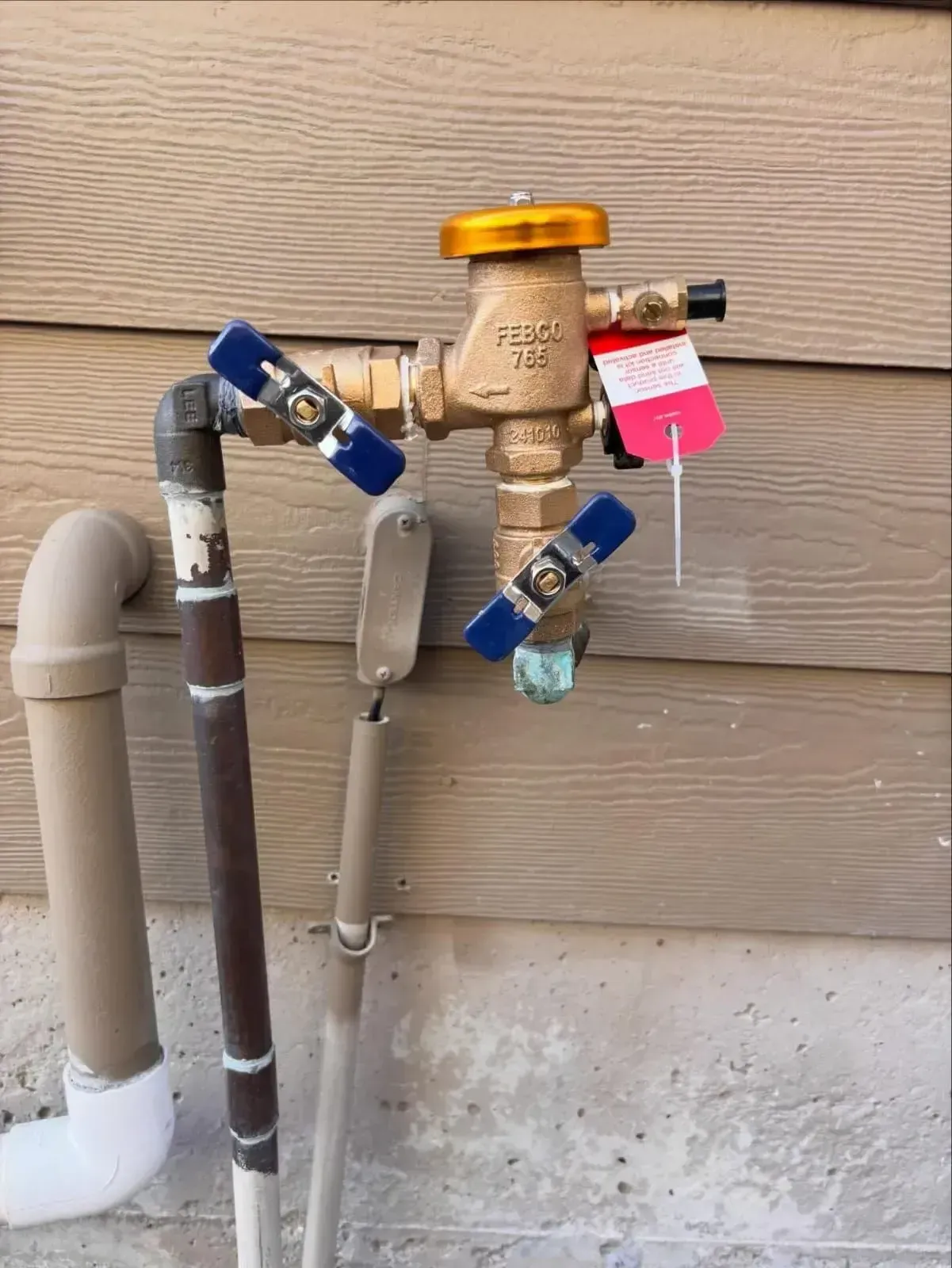 A brass pressure vacuum breaker irrigation valve with blue handles attached to a house exterior wall.