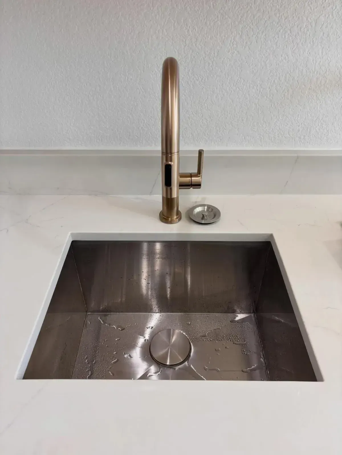 A modern stainless steel kitchen sink set in a white countertop with a copper-tone faucet and a matching drain cover.