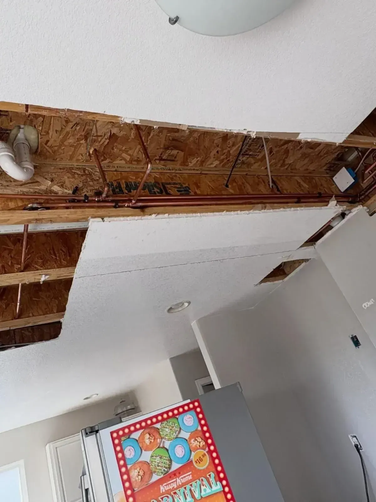 A view looking up at an open ceiling showing exposed wooden joists, copper plumbing pipes, and sections of drywall.