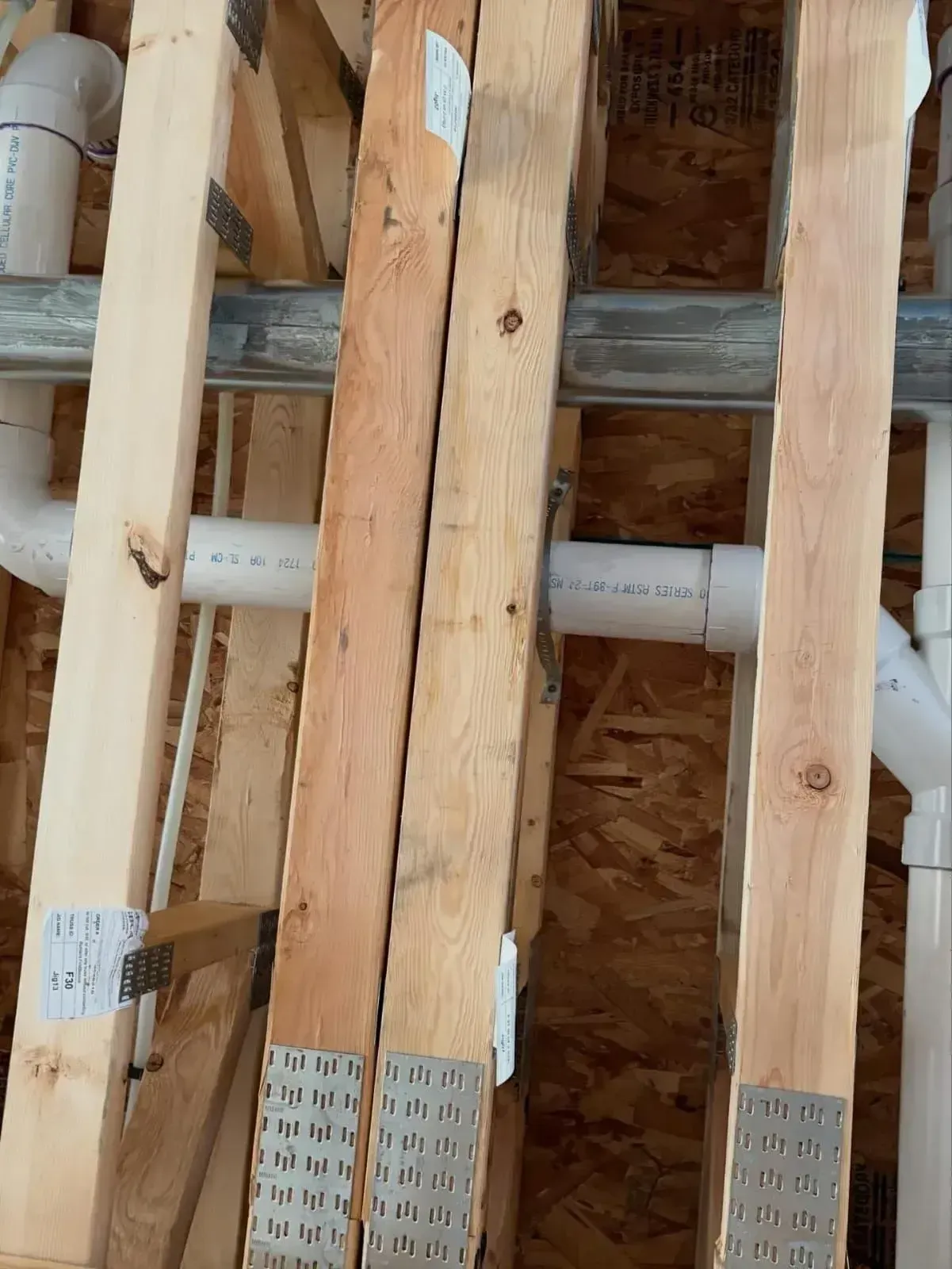 Wooden floor joists with metal connector plates and white PVC plumbing pipes running through them.