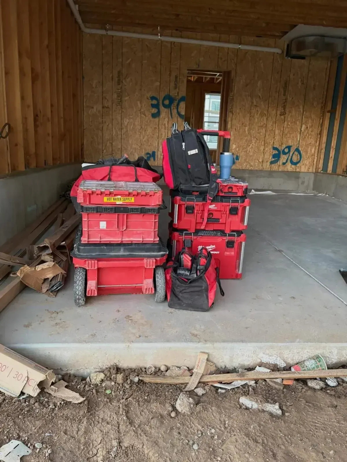 Stacks of red Milwaukee Packout tool storage boxes sitting on a concrete floor in an unfinished wood-framed room.