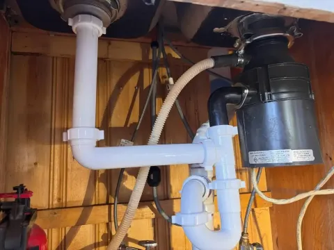 White plastic plumbing pipes connected to a kitchen sink drain and garbage disposal inside a wooden cabinet.