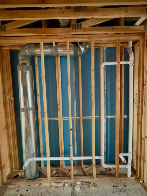 Unfinished wall framing with exposed metal HVAC ducting and white PVC plumbing pipes in a residential construction space.