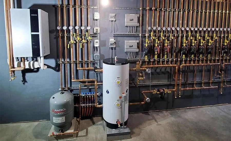 A gray gas water heater sits in a utility closet with a copper piping system and a small expansion tank mounted above.