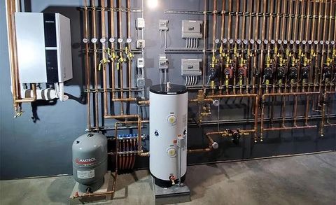 A gray gas water heater sits in a utility closet with a copper piping system and a small expansion tank mounted above.