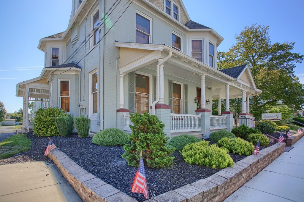 A large white house with a large porch is sitting on the corner of a street.