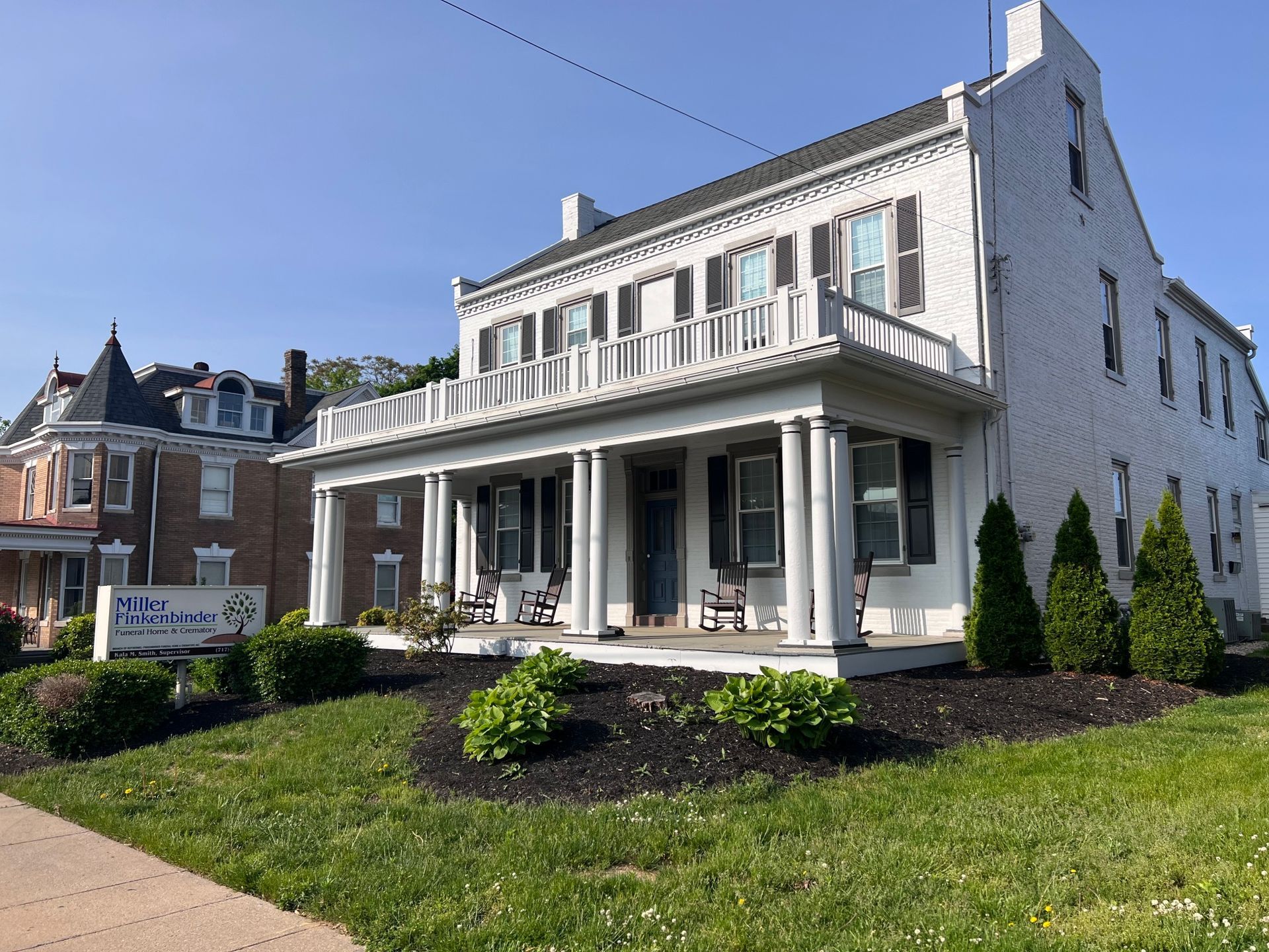 A large white house with a large porch and a brick building in the background.