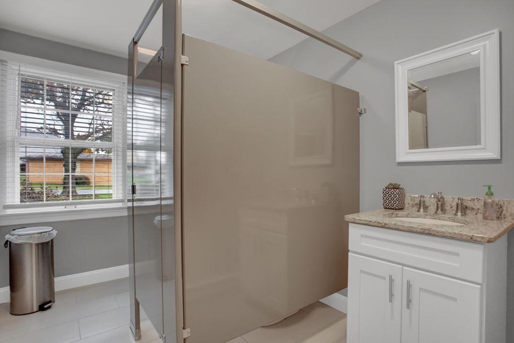 A bathroom with a walk in shower , sink , mirror and trash can.