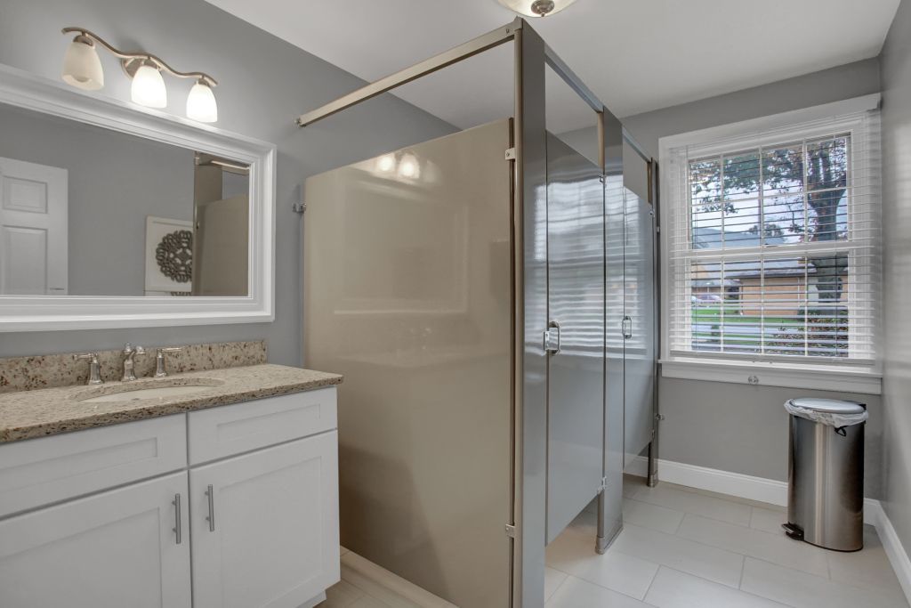 A bathroom with a walk in shower , sink , mirror and trash can.