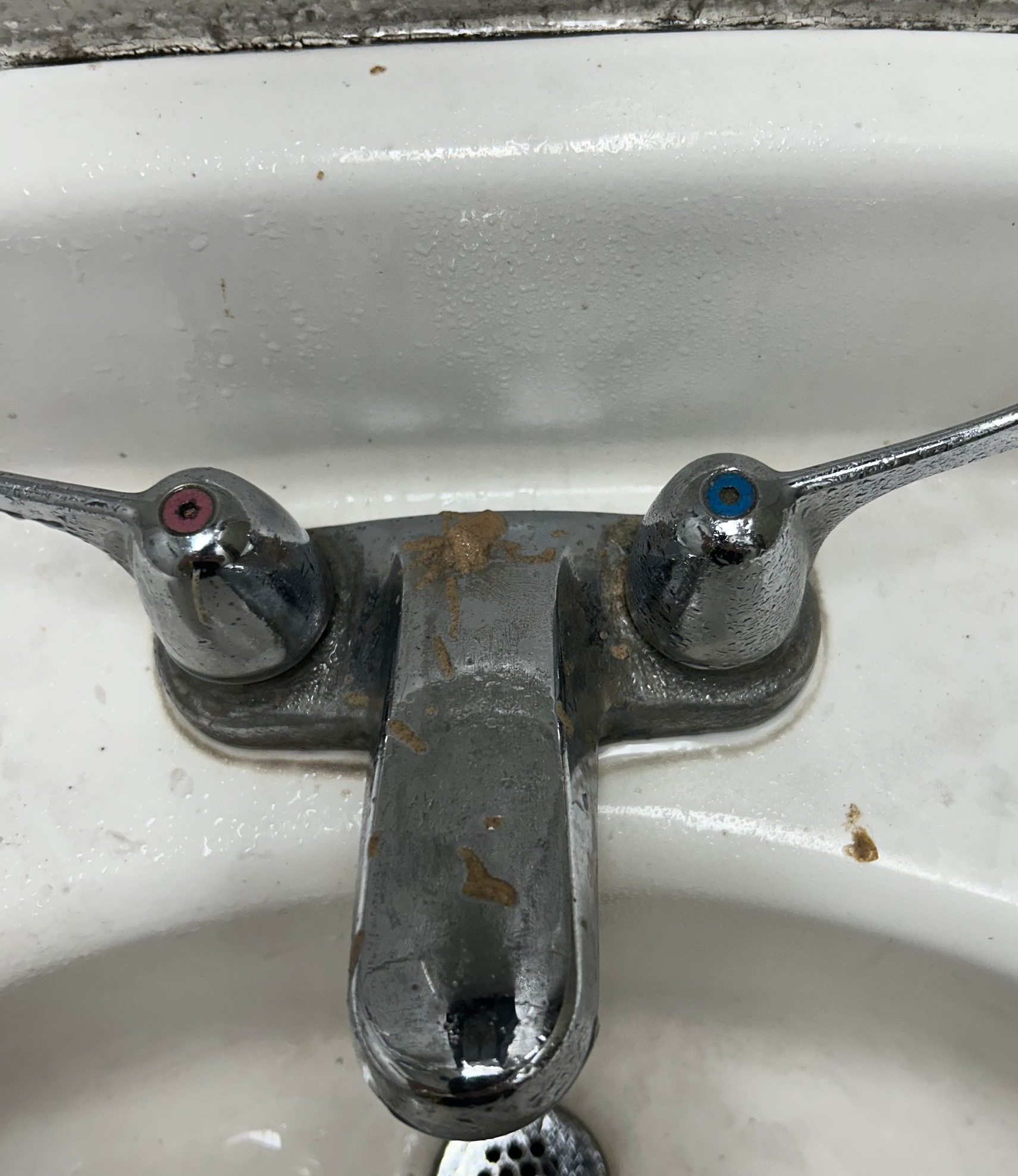 Close-up of a rusty, two-handled faucet in a white sink; one handle is blue, the other is red.