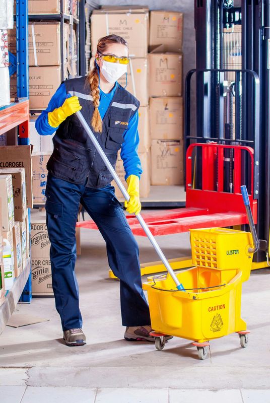 Woman in PPE mopping warehouse floor; boxes in background. Yellow bucket & forklift present.