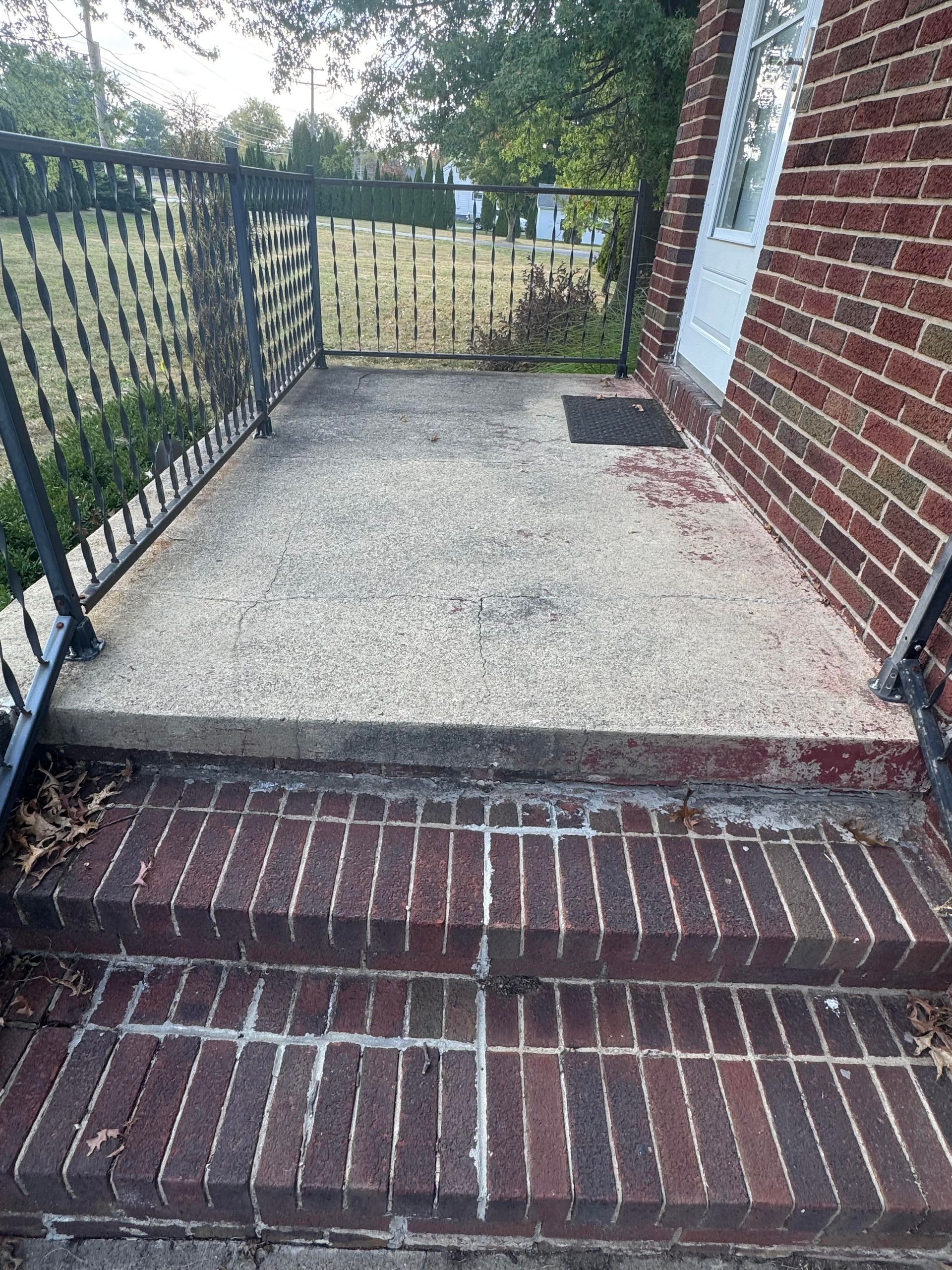 Brick steps leading up to a concrete porch with wrought iron railing and brick wall.