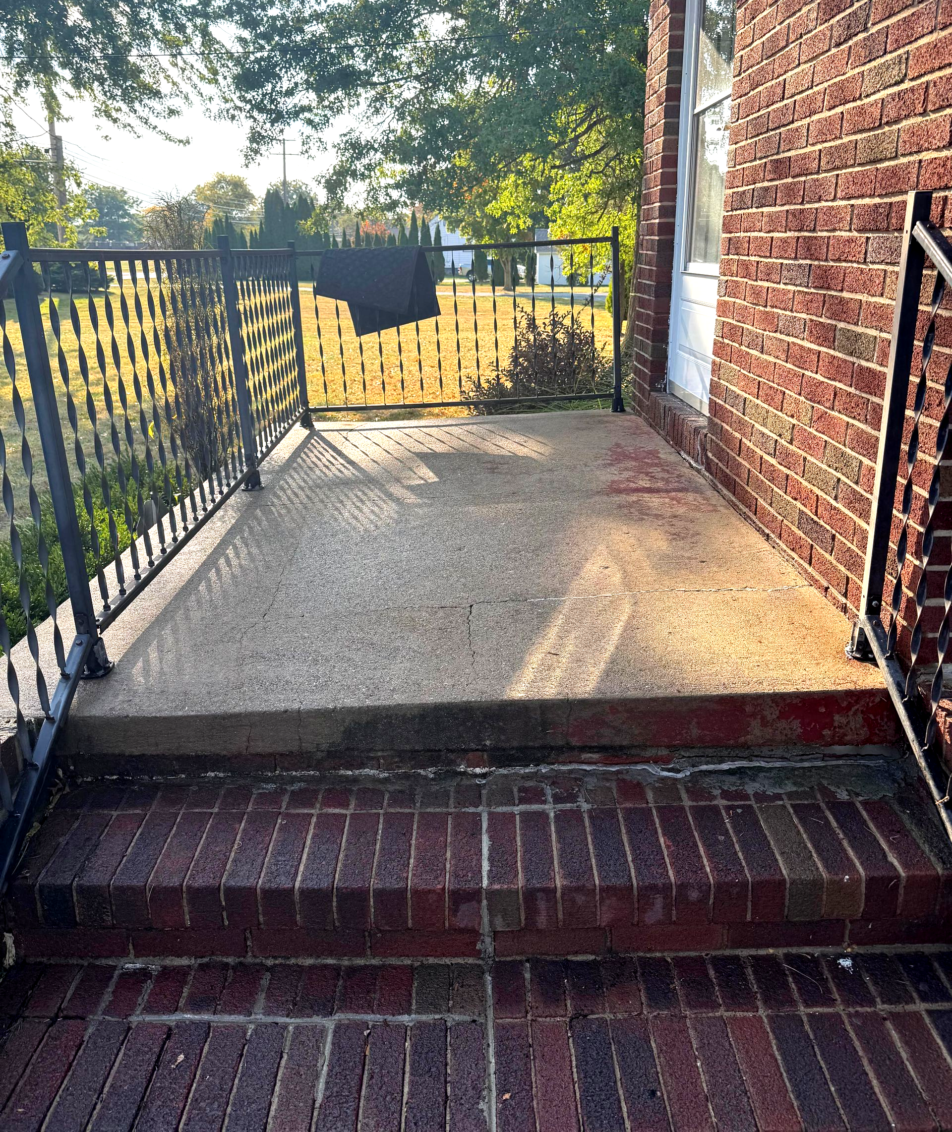 Brick steps lead to a concrete porch with a black wrought-iron railing beside a brick building.