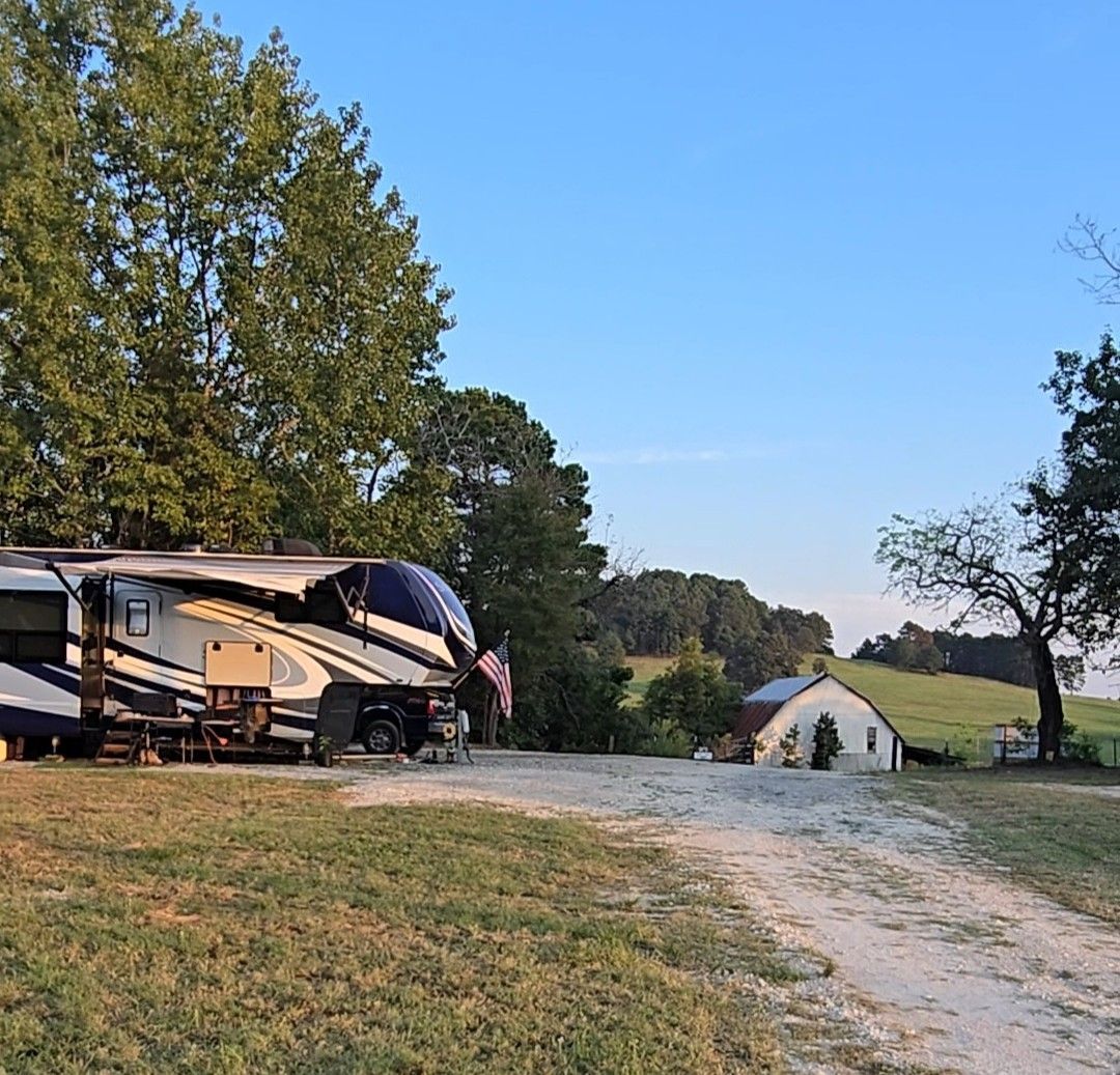 RV parked on grass near a white building and trees under a blue sky.