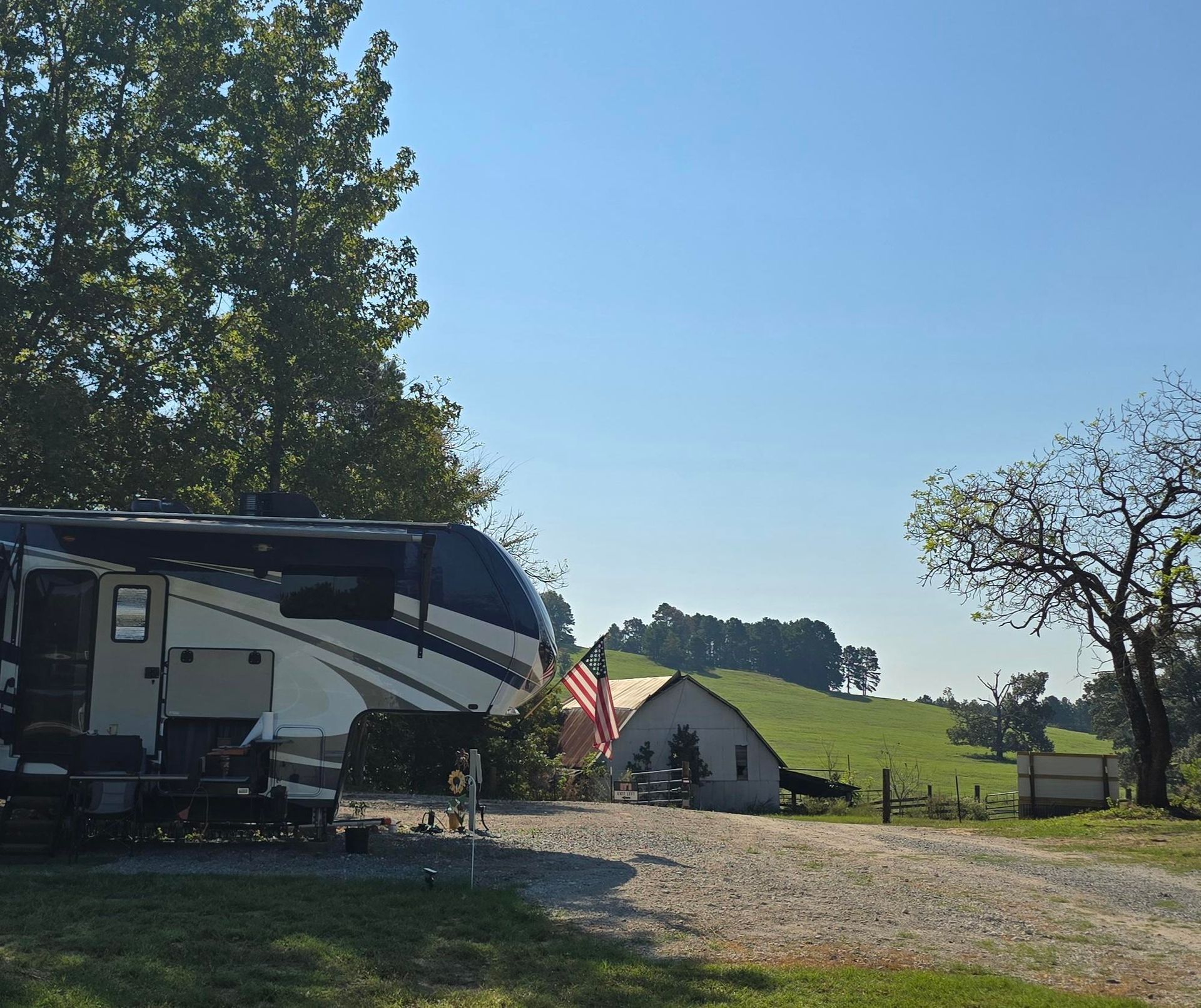 RV parked on a gravel road near a white building and green rolling hills under a blue sky.