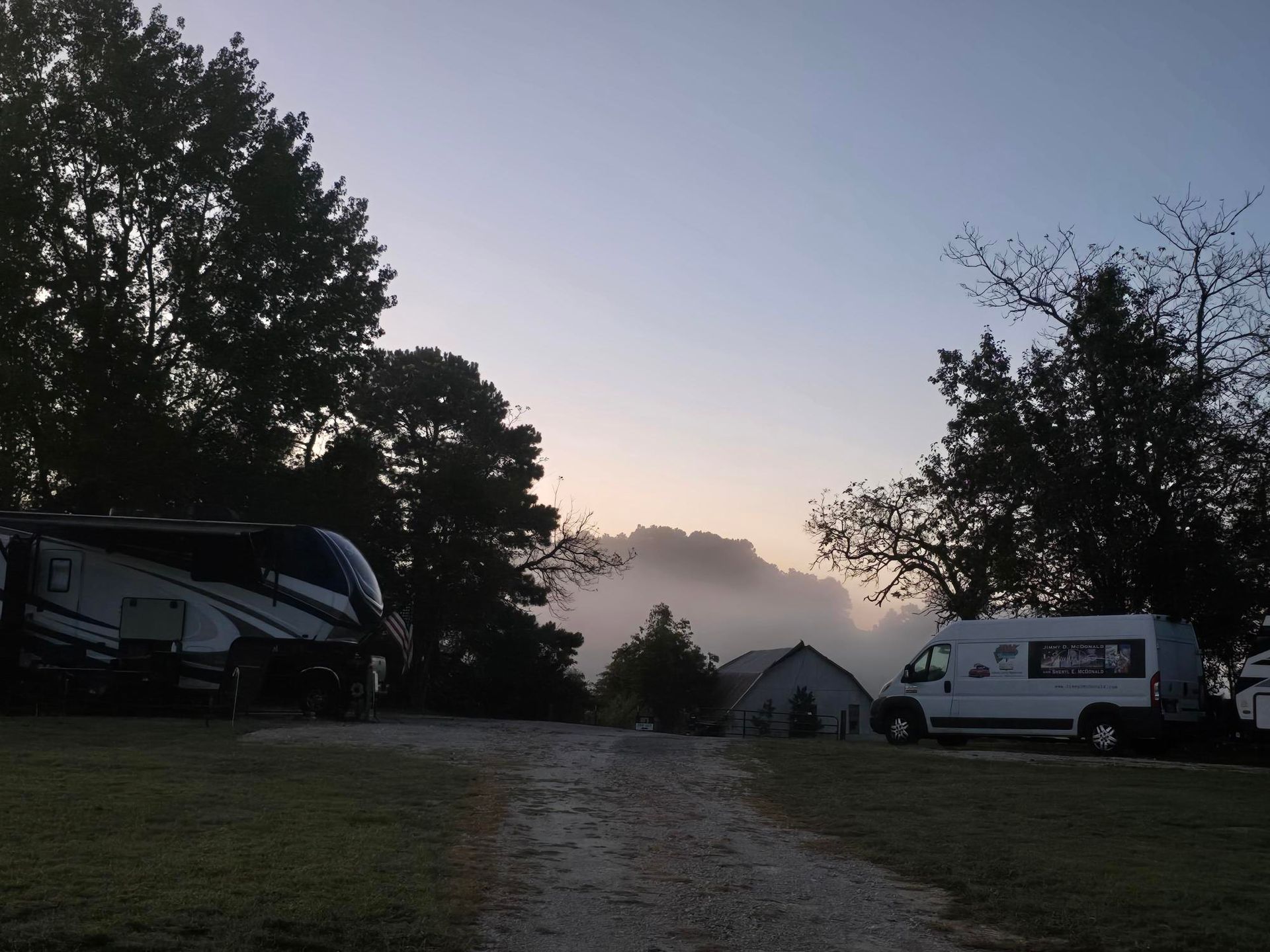RV campers in a misty campsite at dusk. Silhouetted trees surround a gravel path.