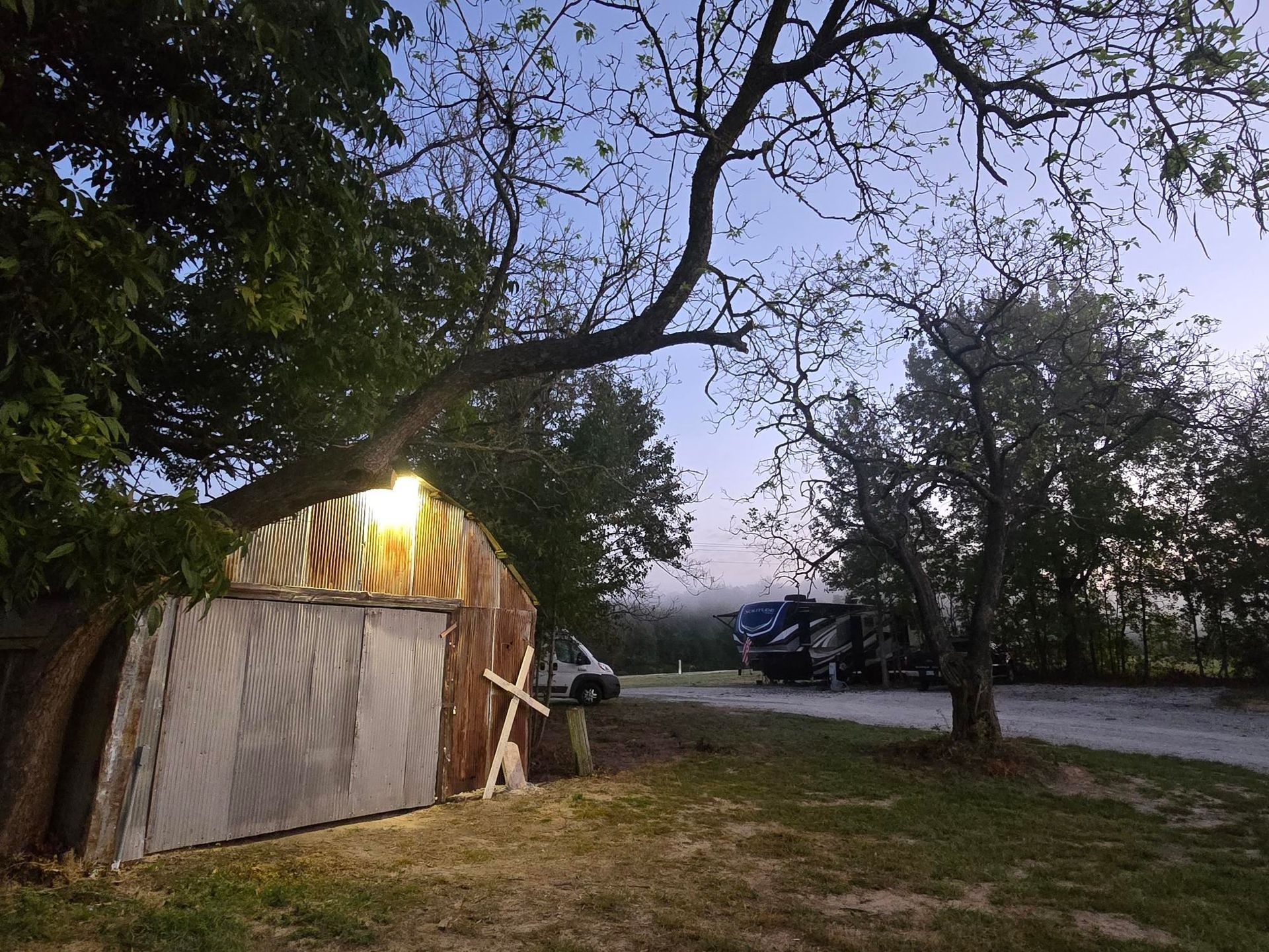 A rustic shed with a light on, trees, RV in a campground at dusk.