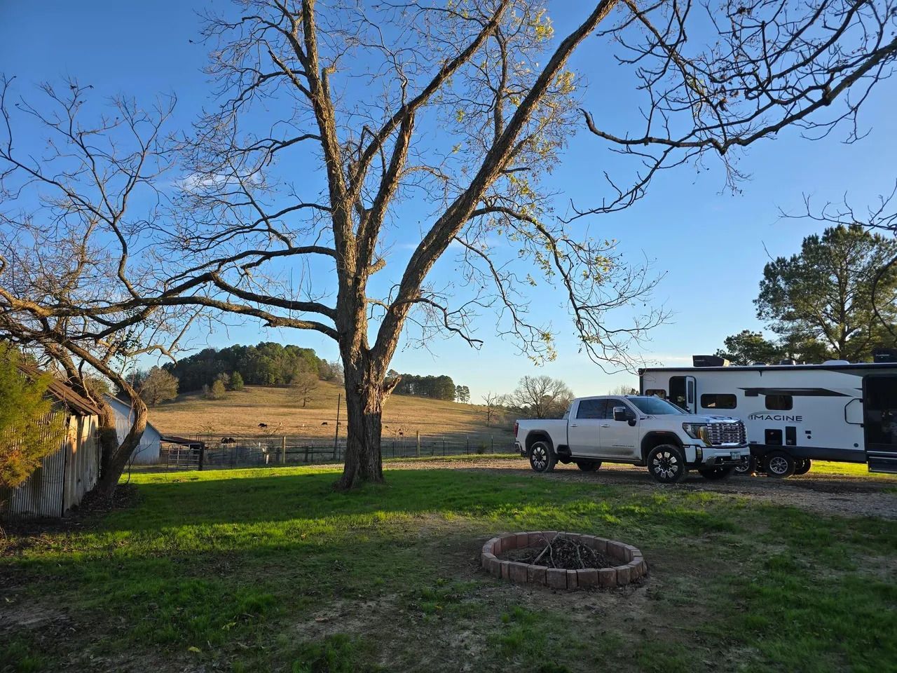 A pickup truck and camper parked on a grassy field near a tree; a fire pit in the foreground.