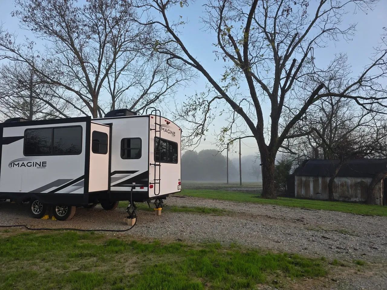 RV parked in a gravel lot next to leafless trees and a small shed, with a misty field in the background.