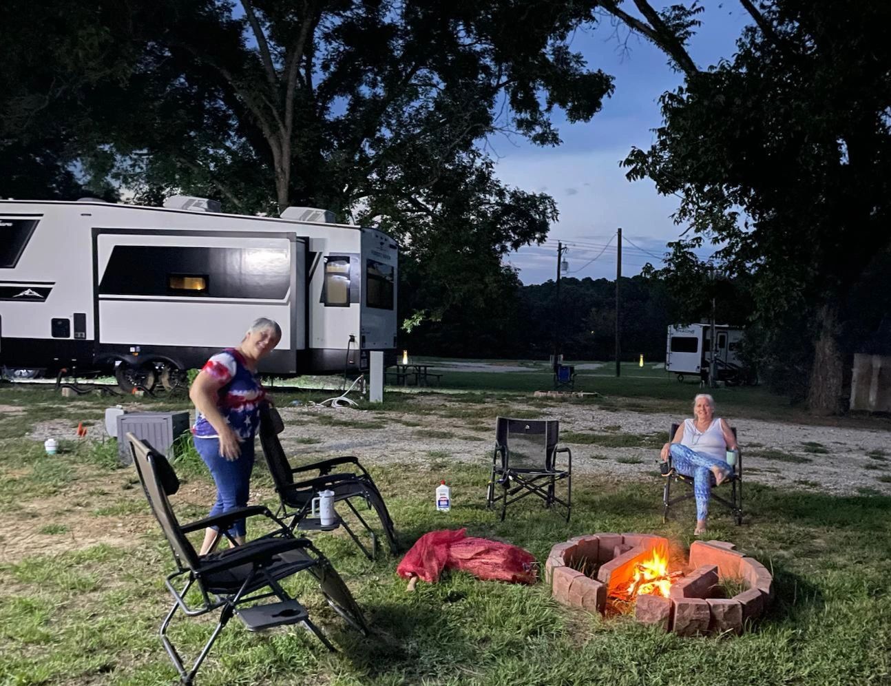 Two women camping at dusk, one by fire, other by RV. Chairs, trees, and RV in setting.
