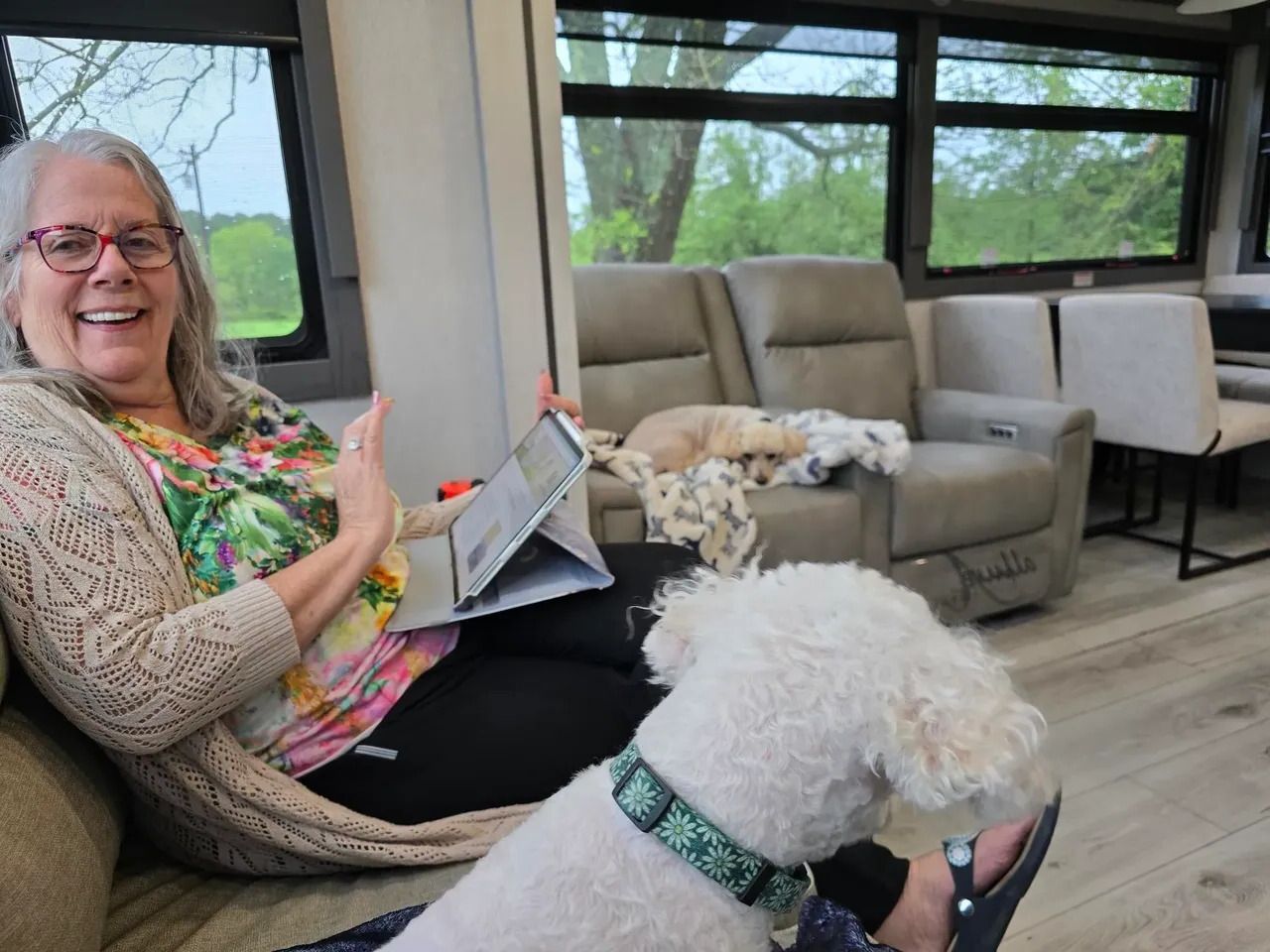 Woman with glasses smiles, holding tablet, and petting a white dog in a modern RV. Another dog rests on a sofa.