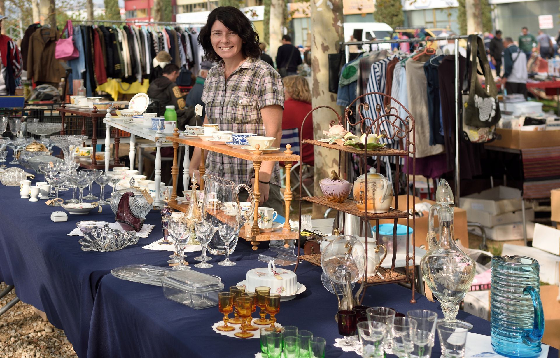 Woman at a flea market stall, selling glassware and clothing. Outdoors, sunny day.