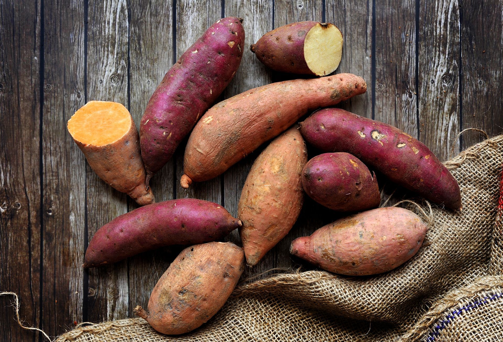 Sweet potatoes, varying in size, color, and shape, rest on a rustic wooden surface with a burlap sack.