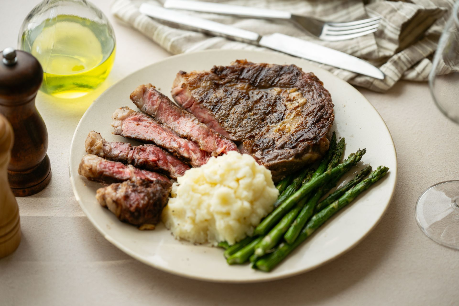 Sliced steak, mashed potatoes, and asparagus on a plate, with utensils, oil, and pepper grinder.