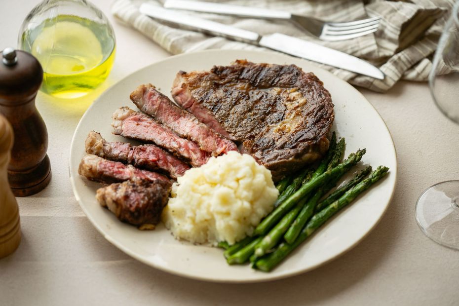 Steak with mashed potatoes and asparagus on a plate, with salt, pepper, oil, knife, and fork.