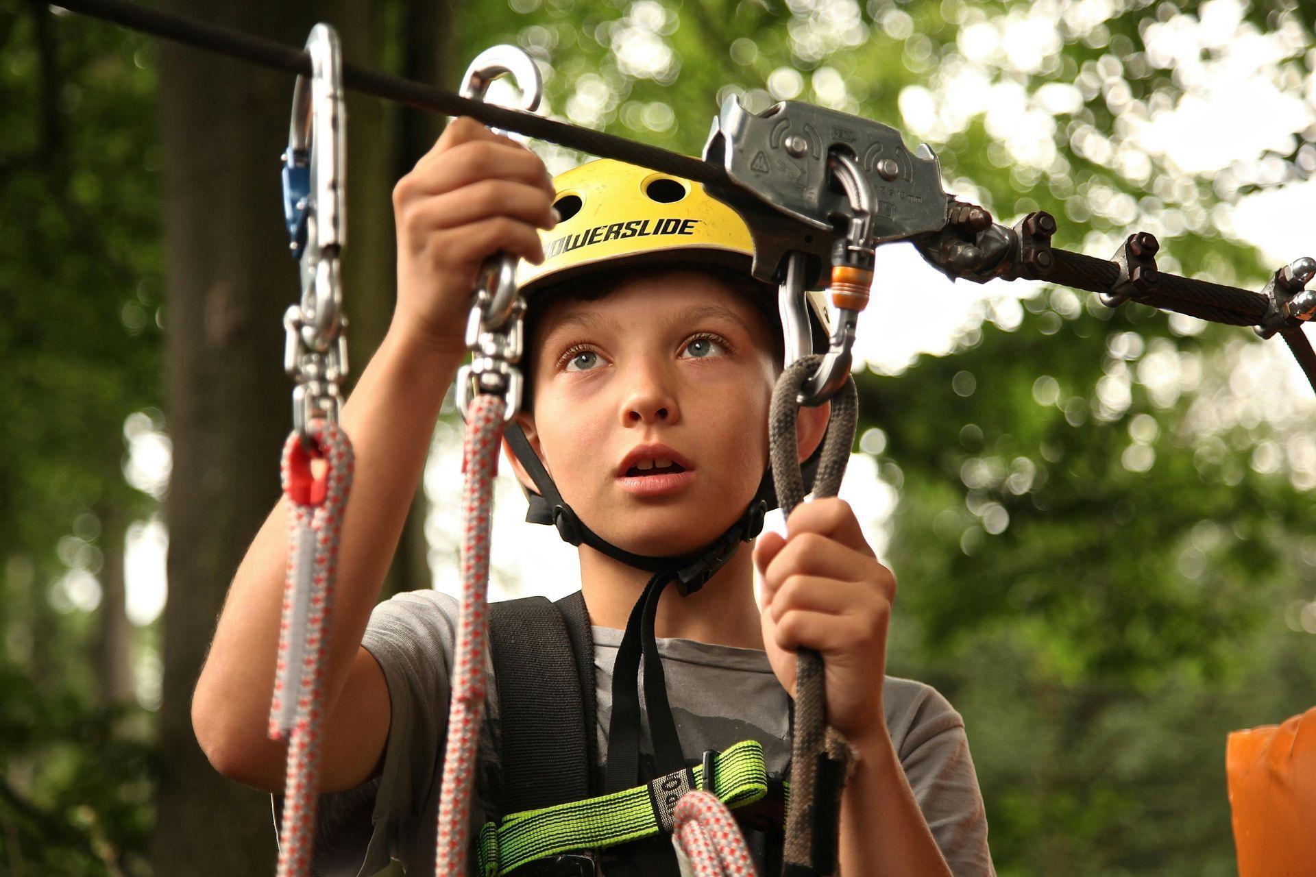 Boy in a helmet on a zip line course, looking upward, holding safety gear.