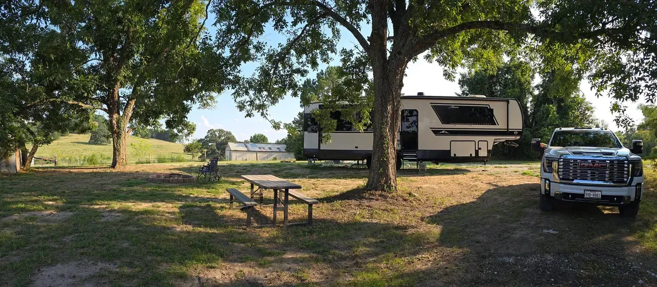 A camper and truck parked at a campsite under a large tree, next to a picnic table and green field.