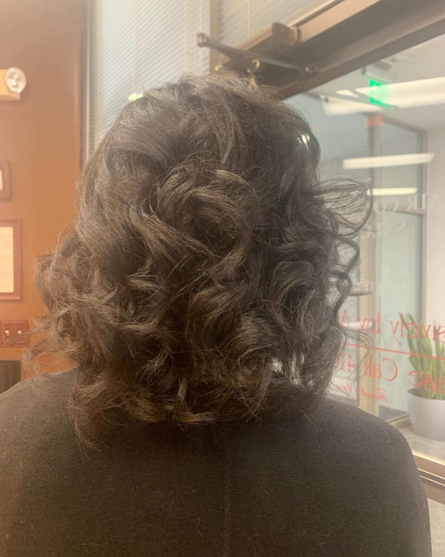 Person with short, dark, curly hair; back view. Standing indoors near a window and wall.