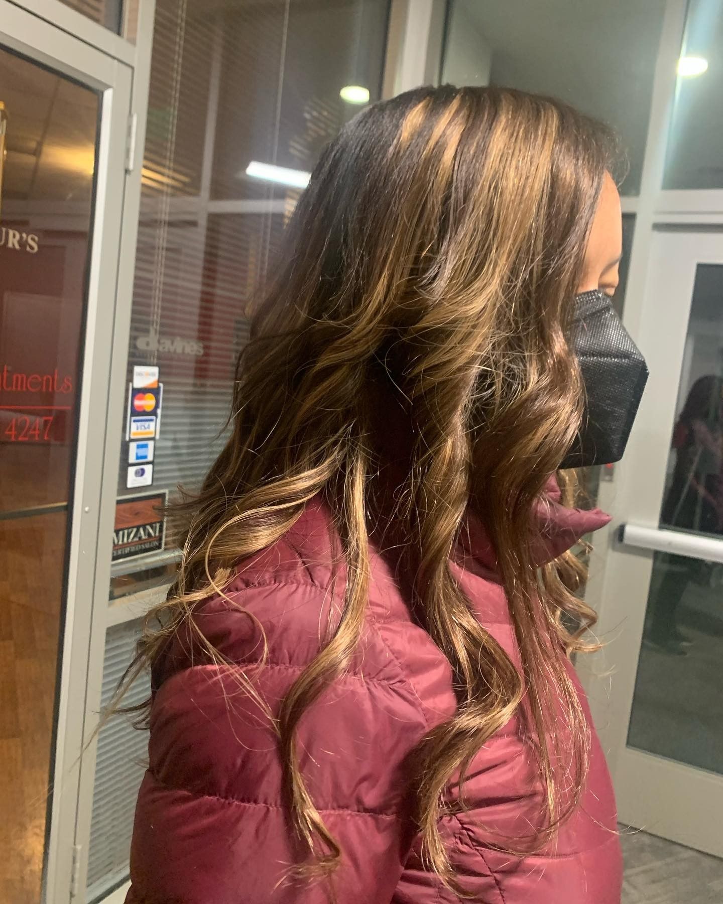 Woman with long, wavy brown hair, wearing a black face mask and a burgundy coat, standing outside a building.