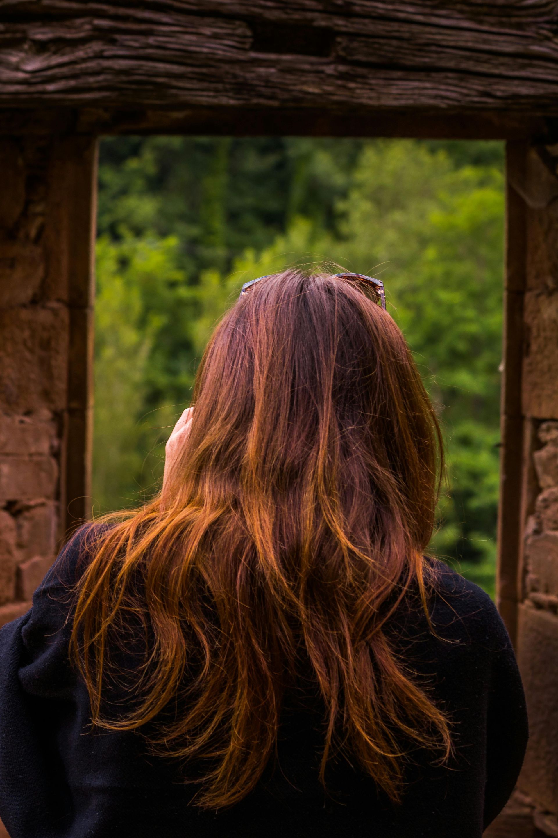 Woman with long red hair looking out window at green trees.