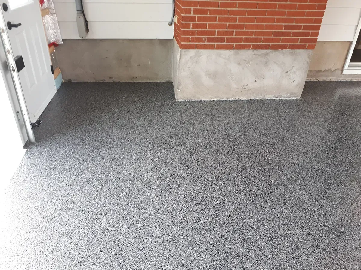 A floor with a gray and black speckled epoxy finish next to a white door and a brick and concrete foundation wall.