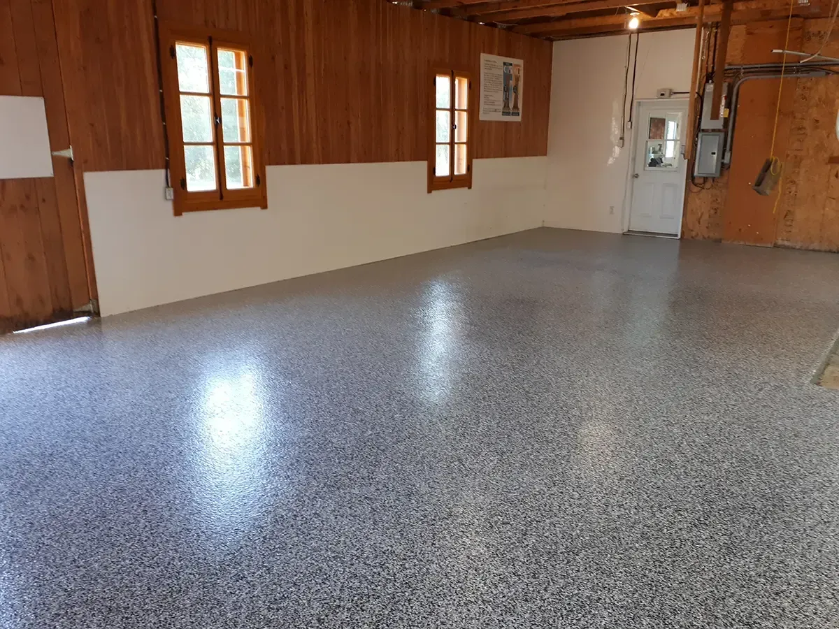 Interior of a room with wood-paneled walls, white trim, two windows, and a shiny, speckled grey epoxy floor.