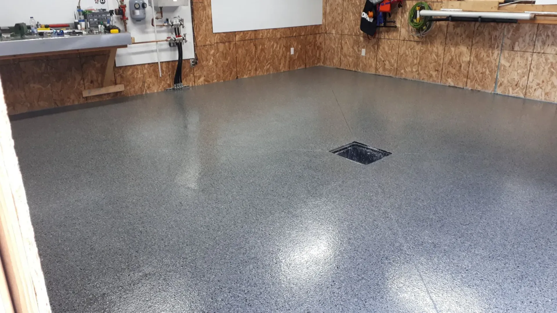 A freshly coated gray speckled garage floor with a drain cover, situated between workbench areas and wood-paneled walls.