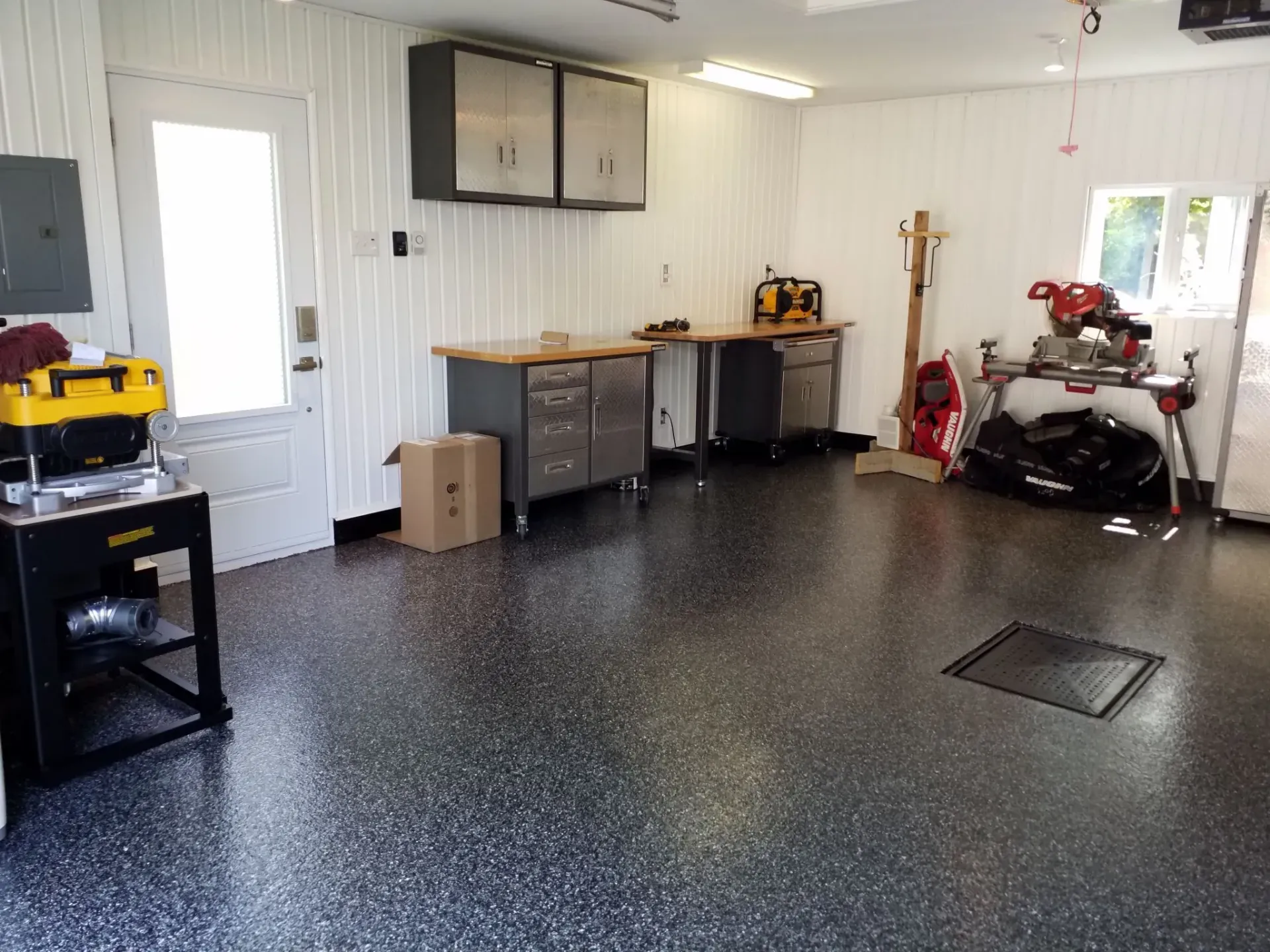 Garage interior with workbenches, tools, cabinets, and a shiny, speckled floor. White walls, door, and natural light.