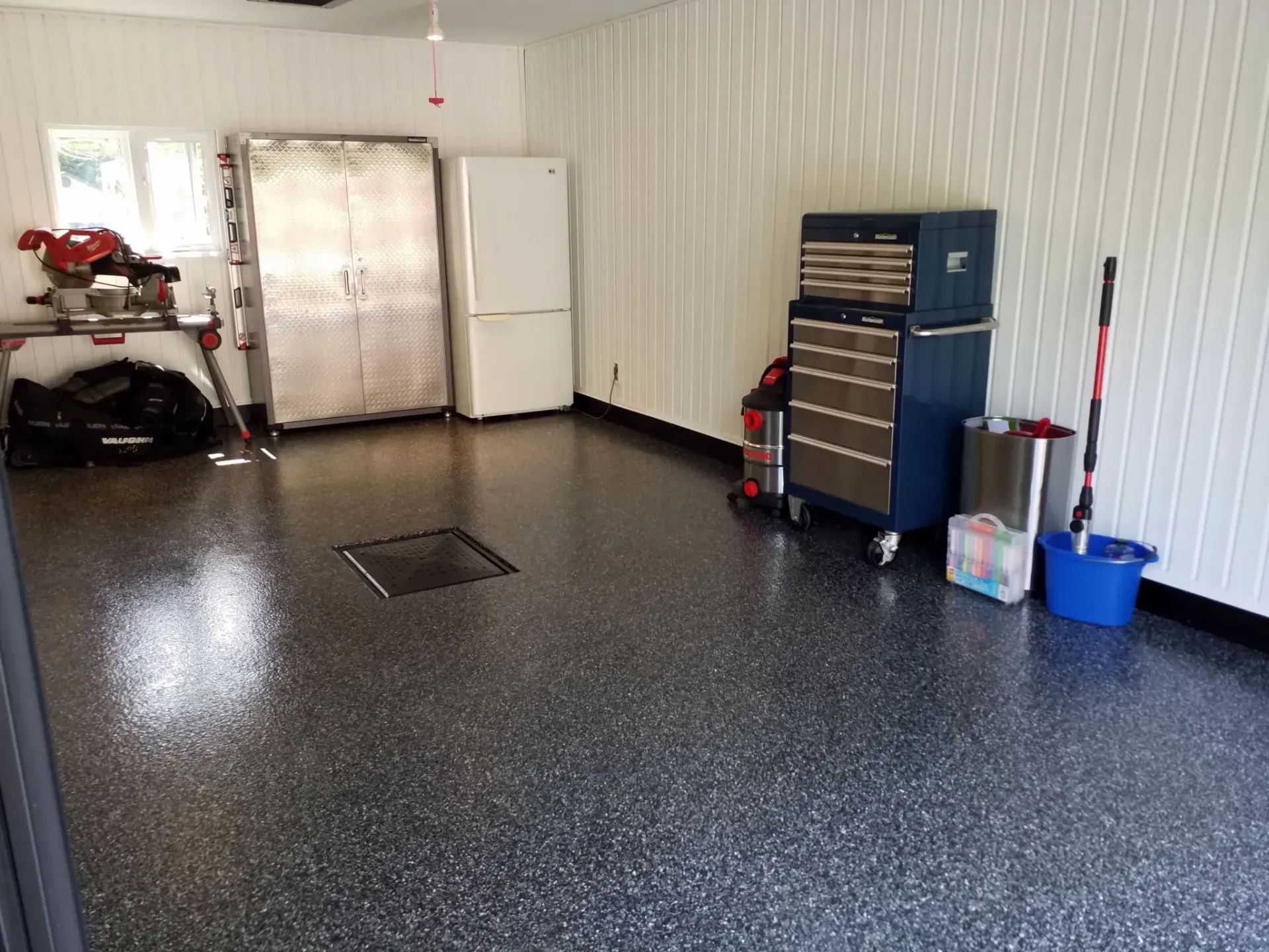 A clean garage with a black and white speckled epoxy floor, white panel walls, a metal cabinet, fridge, and tool chest.