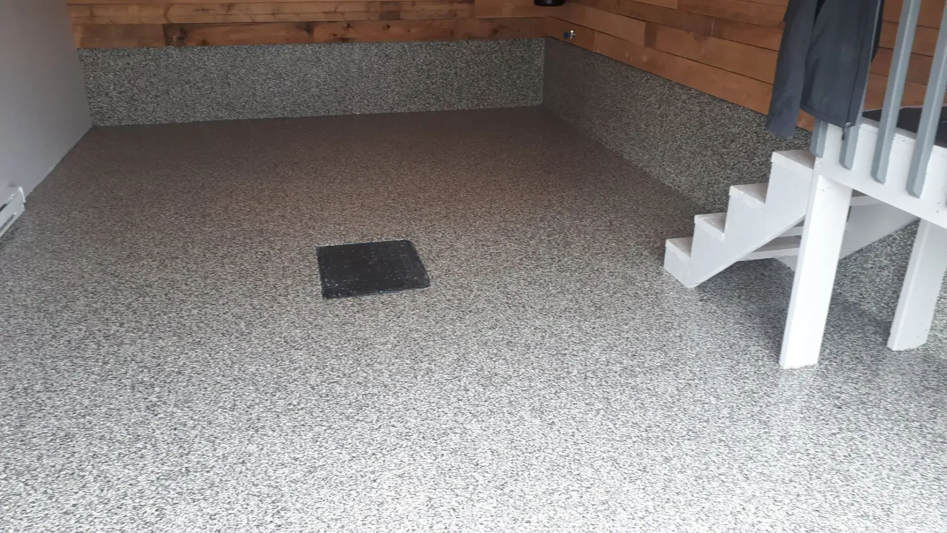 Garage with speckled gray flooring, dark square drain, white stairs and wood paneling on the wall.