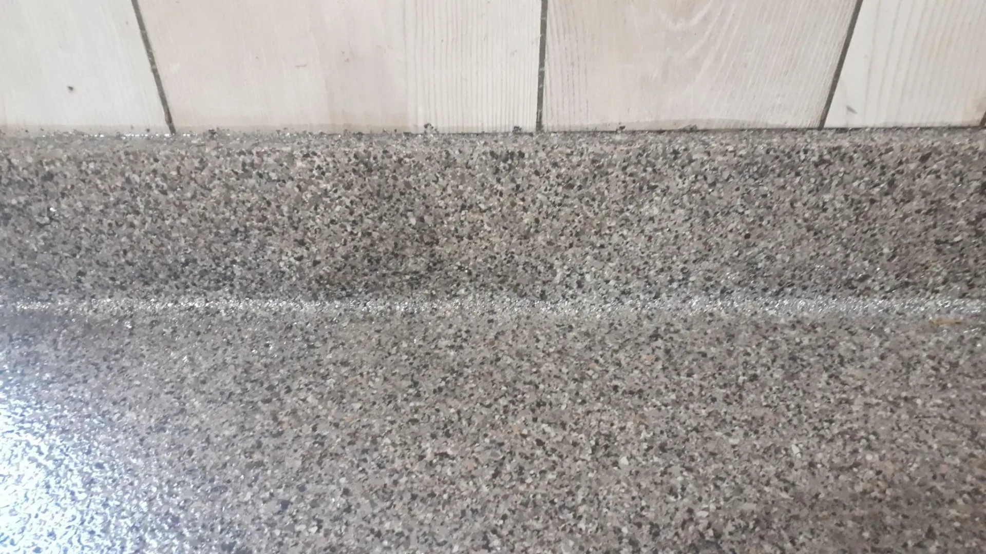 Close-up of a speckled, textured, grey-brown flooring next to light wood panels.