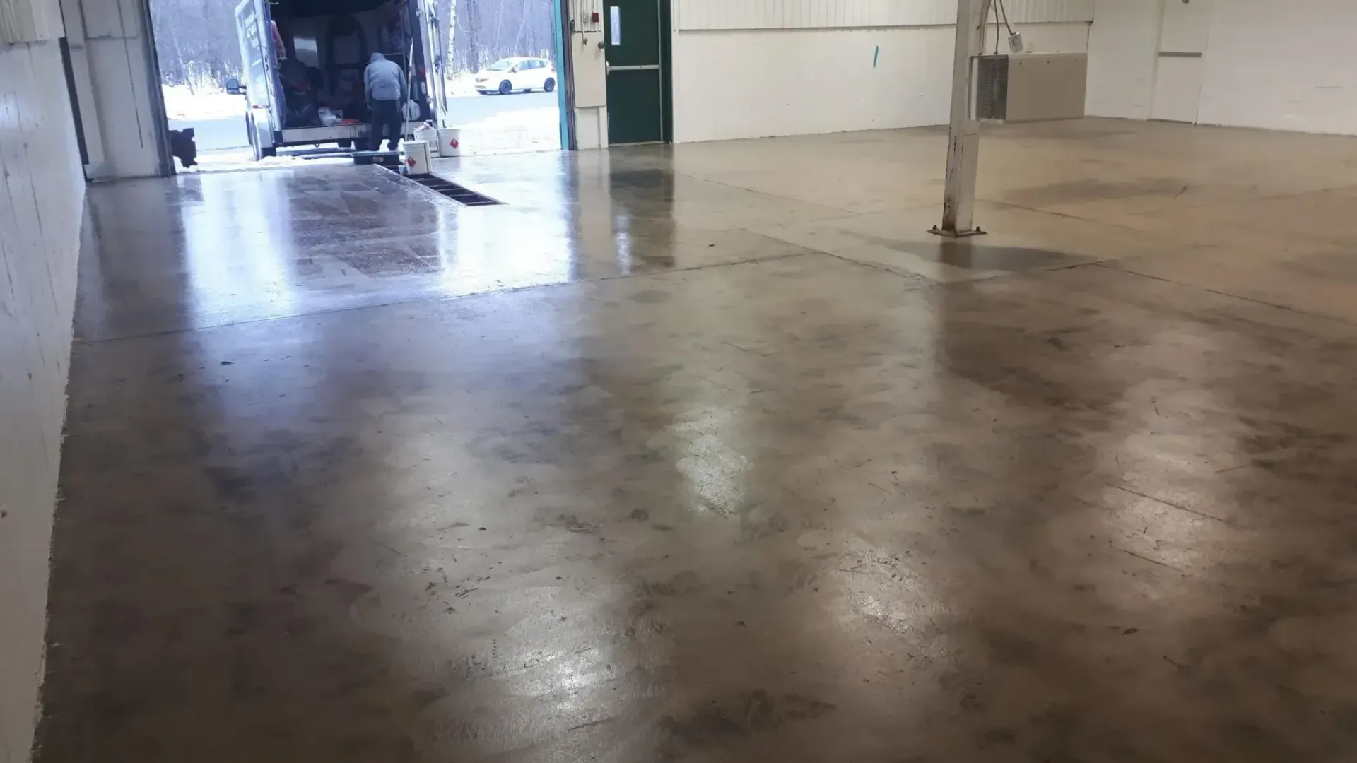 Inside industrial space, wet concrete floor reflects light. A vehicle is parked in an open doorway.
