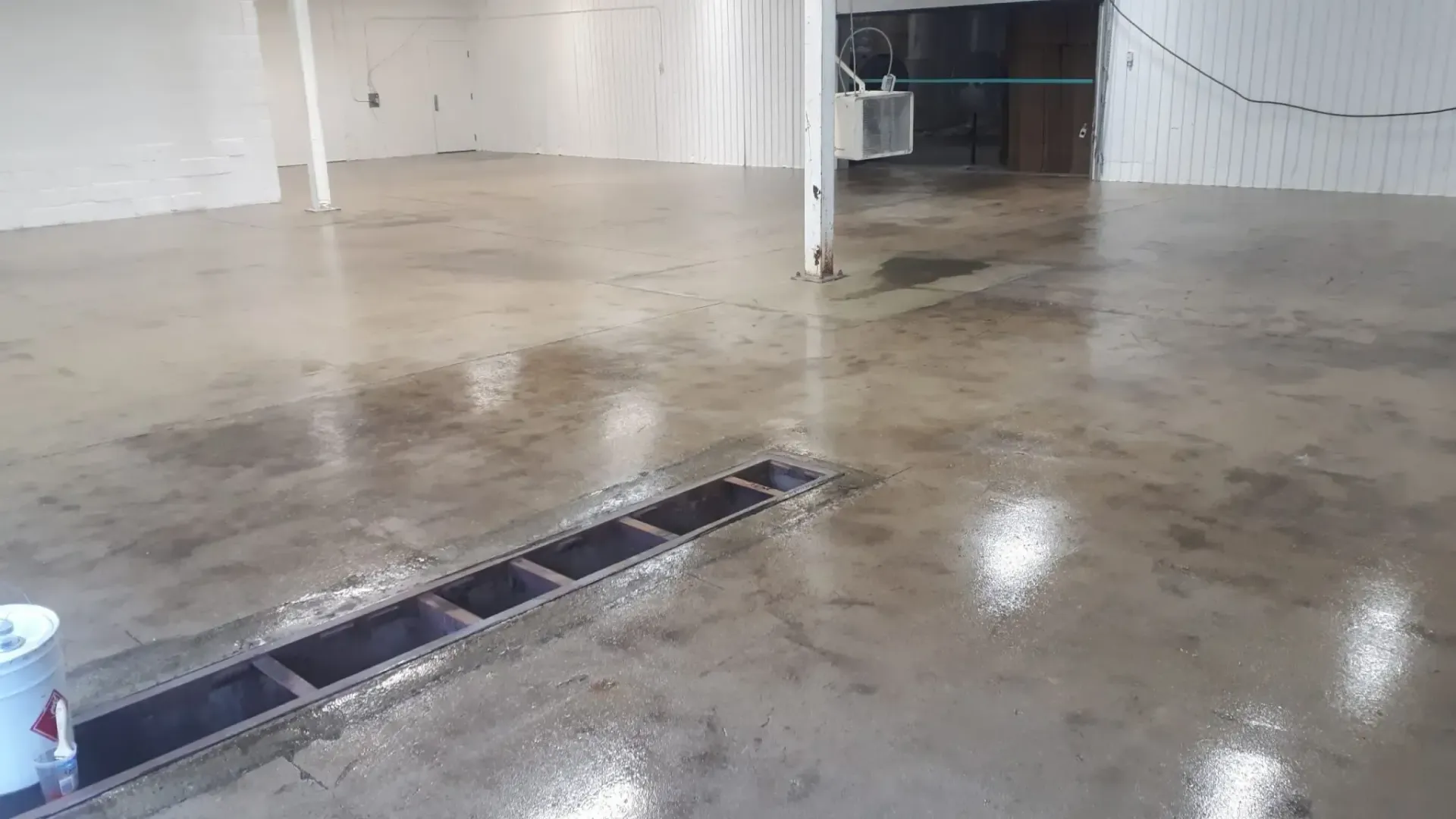 A wet, gray concrete floor with a recessed drain. White walls and columns in an empty room.