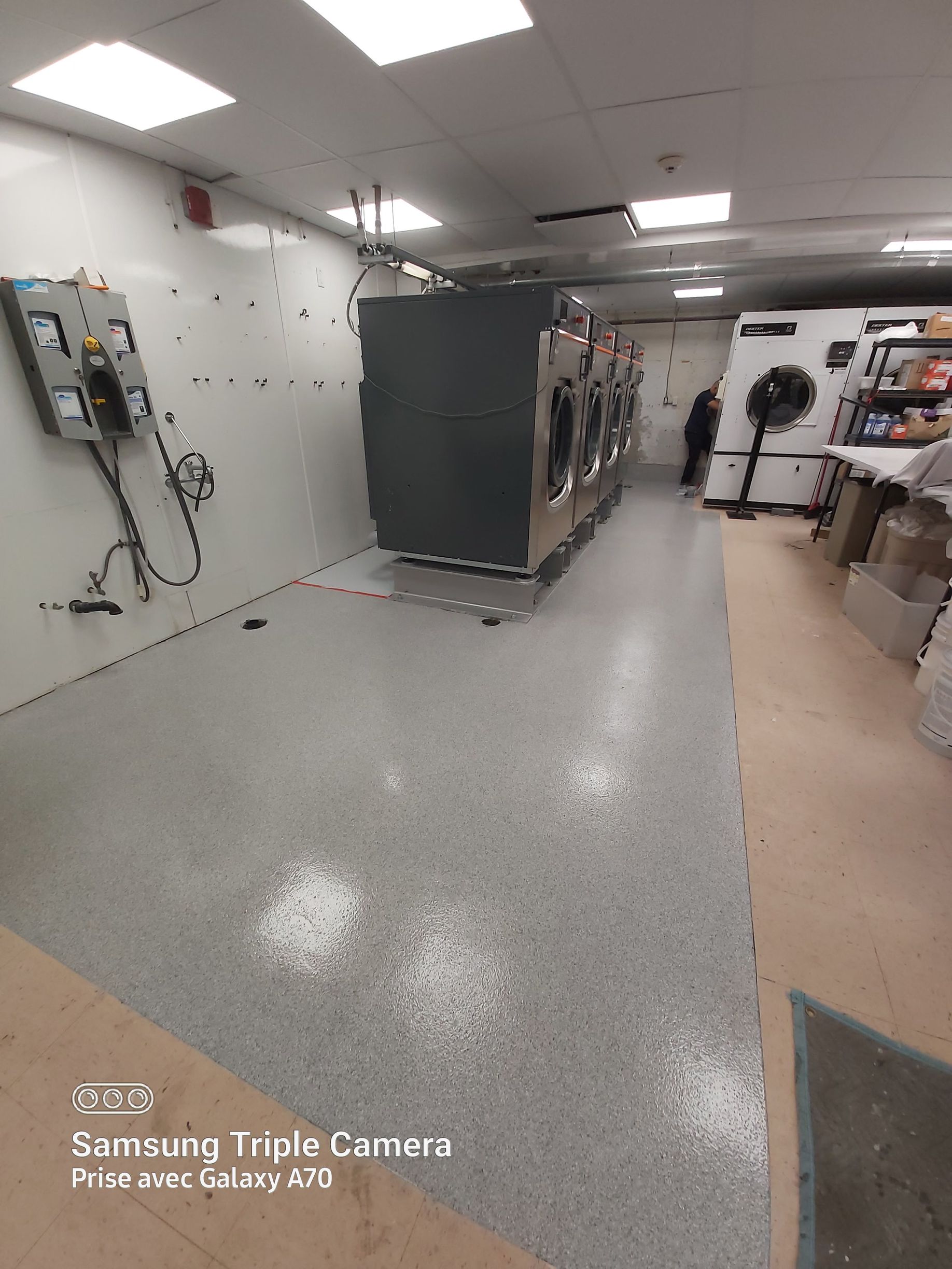 Laundry room with industrial dryers. Gray epoxy floor, white walls, and a person in the background.