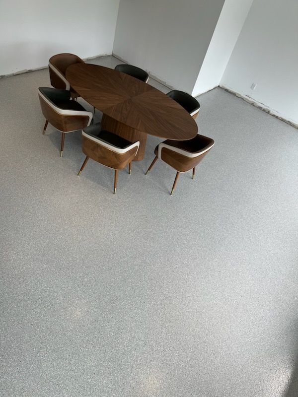 Oval wooden table with six chairs on a speckled grey floor in a mostly empty room.
