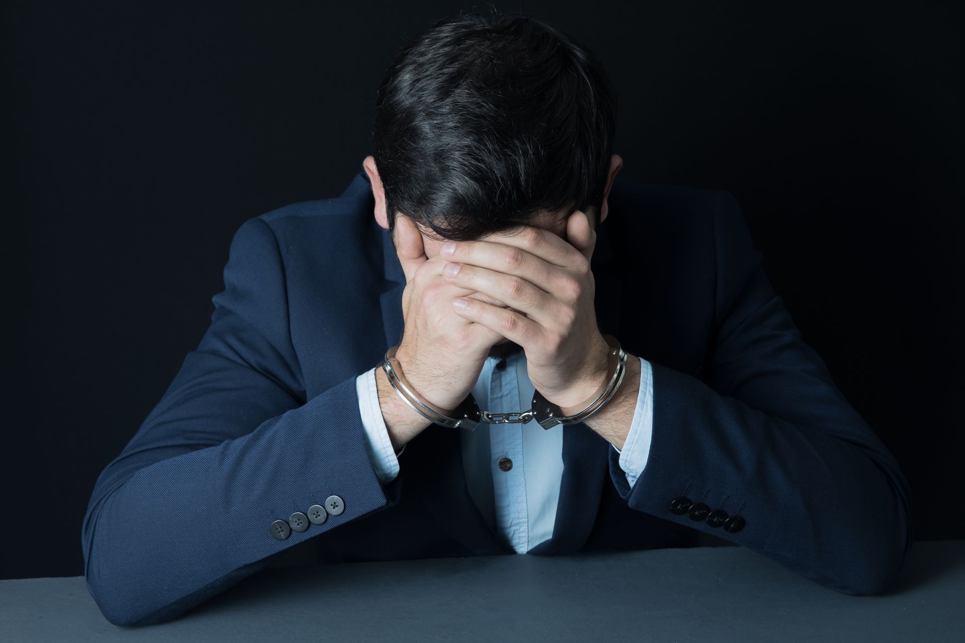 Man in suit, handcuffed, head in hands, appears distressed.